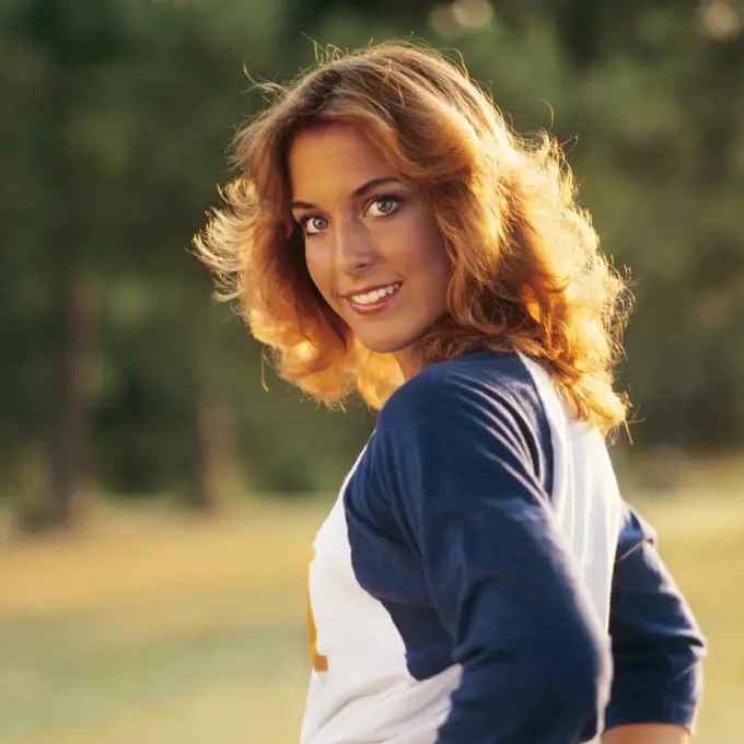 1970s 1980s SMILING YOUNG WOMAN SUNLIGHT ON HER GOLDEN HAIR LOOKING AT CAMERA OVER HER SHOULDER