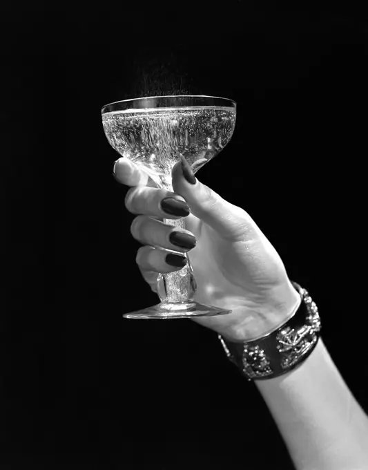 1930S 1940S 1950S Woman Hand Ornate Metal Bracelet Holding Up New Year Toast Glass Of Champagne Against Black Background