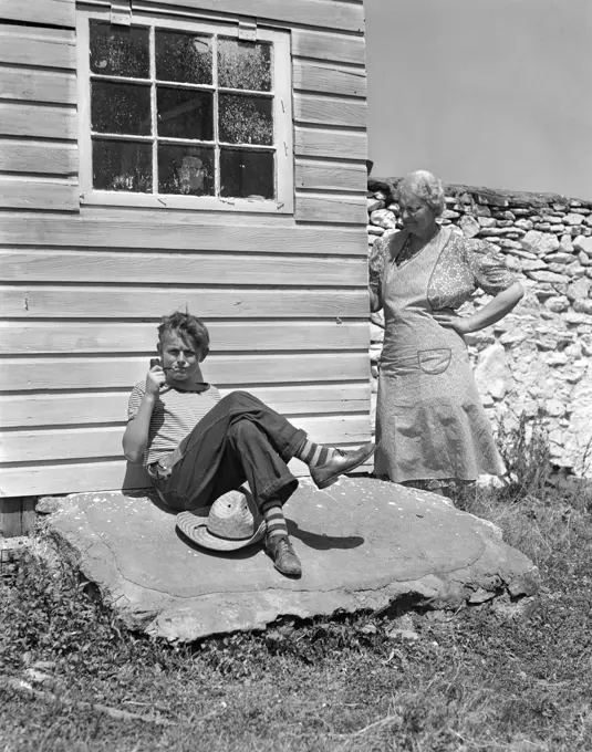 1940S Farm Boy Leaning Against House Smoking Pipe With Grandmother Looking Around Corner At Him With Hand On Hip