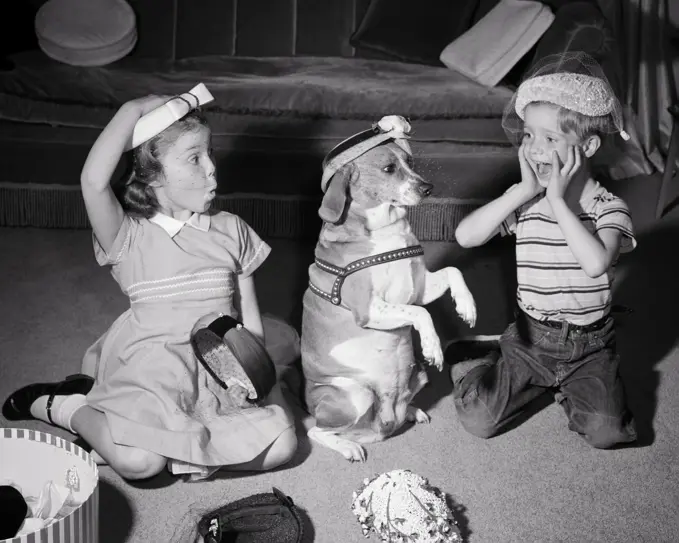 1950s SISTER AND BROTHER PLAYING WITH AND WEARING MOTHERS HATS AND PUTTING ONE ON BEAGLE DOG