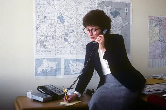 1980s WOMAN REALTOR SITTING ON DESK TALKING ON TELEPHONE AND WRITING A MESSAGE MAPS ON THE WALL