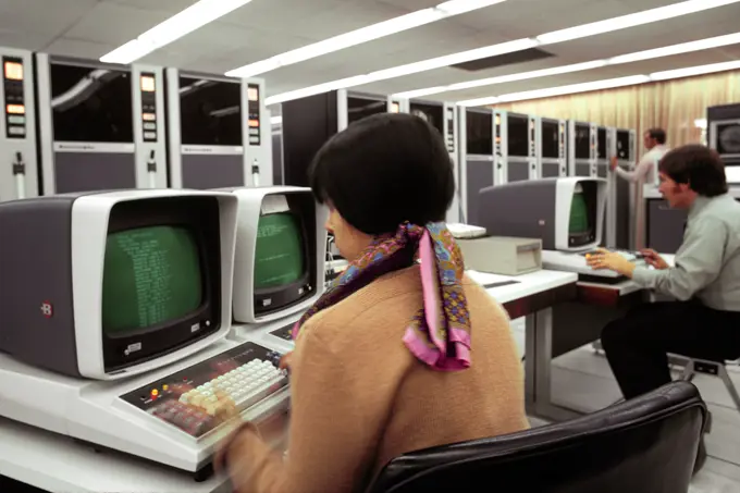 1970s YOUNG WOMAN TECHNICIAN SITTING IN FRONT OF BURROUGHS COMPUTER USING KEYBOARD IN BACKGROUND ARE MAGNETIC TAPE TRANSPORTS