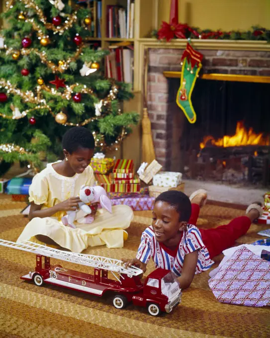 1970s AFRICAN-AMERICAN BROTHER AND SISTER BY CHRISTMAS TREE AND FIREPLACE GIRL HOLD STUFFED DOG TOY AND BOY WITH TOY FIRE TRUCK