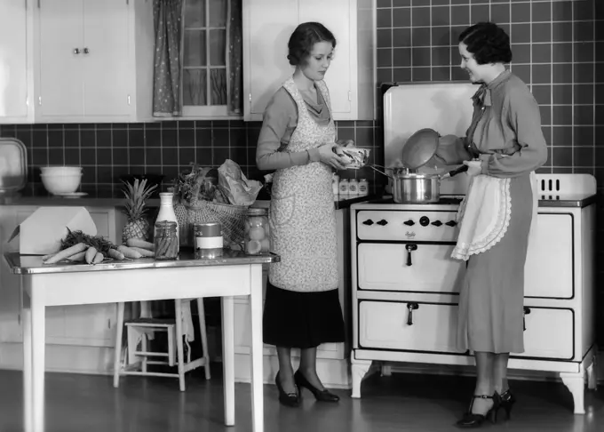 1930S Woman Housewife And Friend Wearing Apron Cooking Food In Kitchen On Gas Stove Indoor