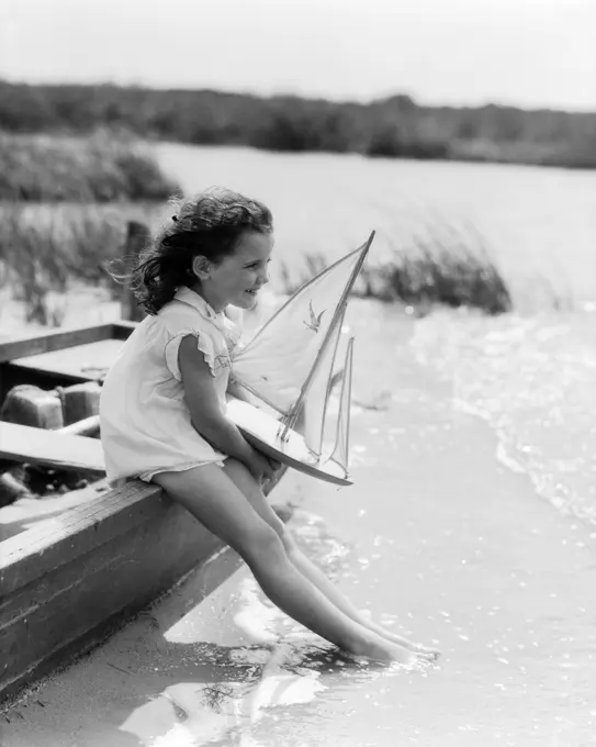 1930S Young Girl At Seashore Holding Sailboat Toy Sitting On Edge Of Rowboat Feet In Water