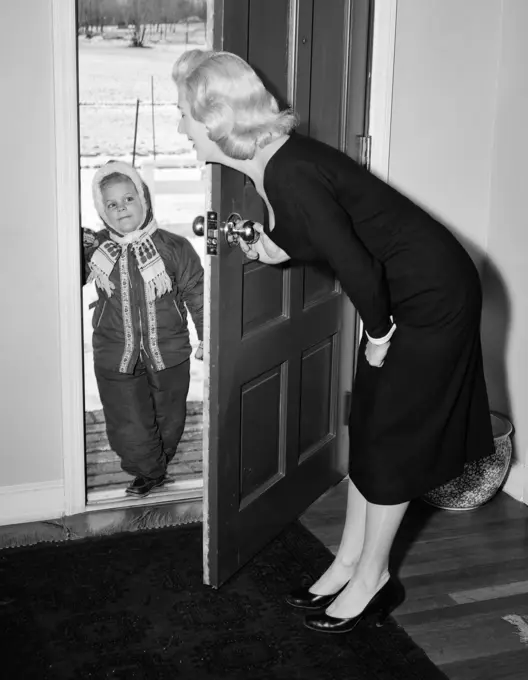 1950S Blonde Mom Bending Opening Front Door To Let In Child Dressed In Winter Snow Suit Scarf Hat Can See Snow Scene Through Door Come In Get Warm