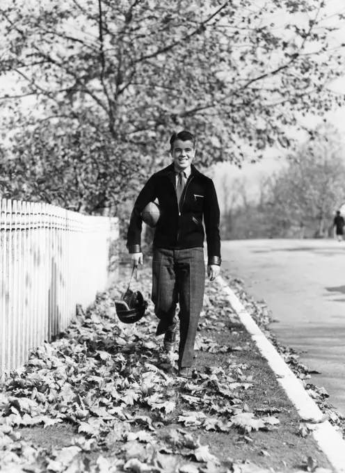 1930S 1940S Smiling Boy Walking Autumn Street Home From School Carrying Helmet And Football