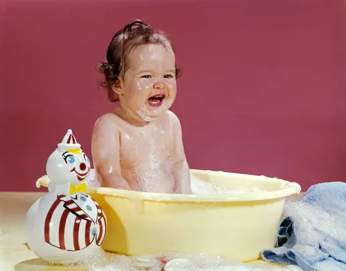 1960S Crying Brunette Baby Sitting In Yellow Plastic Bath Tub With Toy Clown