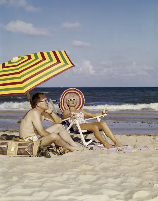 1950S 1960S Middle Age Couple Sitting On Beach Under Umbrella Man With Golf Clubs Looks Bored Wife Happy