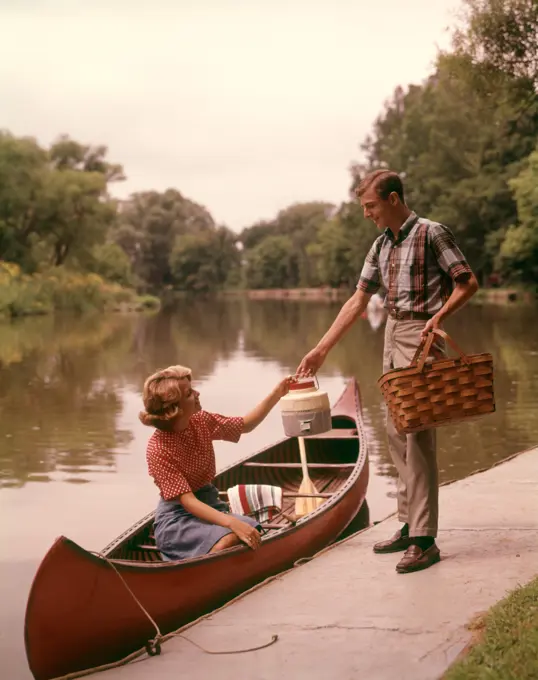 1960S Young Couple Loading Picnic Basket Thermos Into Canoe  