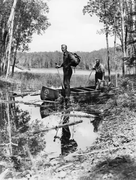 1930s 1940s TWO MEN STANDING IN CANOE IN SHALLOW WATER OF LAKE HOLDING OARS WEAR BACKPACKS LAKE OF THE WOODS ONTARIO CANADA