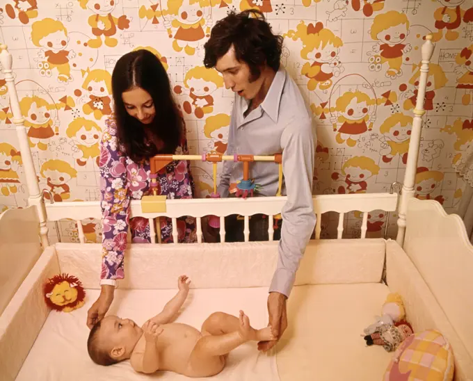 1970s MOTHER FATHER COUPLE LOOKING TOUCHING BABY LYING IN NURSERY CRIB