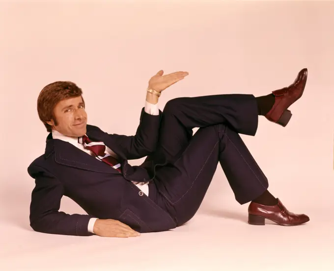 1970s SMILING MAN IN BUSINESS SUIT AND TIE LYING DOWN LEGS CROSSED LOOKING AT CAMERA MAKING UPLIFTING GESTURE WITH HAND