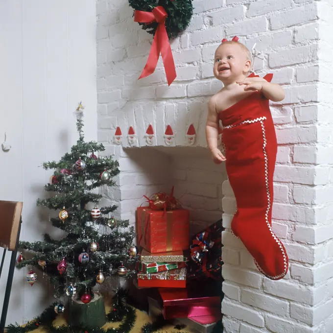 1960s BABY GIRL STUFFED IN CHRISTMAS STOCKING HANGING NEAR FIREPLACE SURROUNDED BY PRESENTS AND TREE