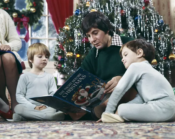 1970s FATHER READING NIGHT BEFORE CHRISTMAS STORY TO 2 BOYS BY TREE SITTING ON FLOOR