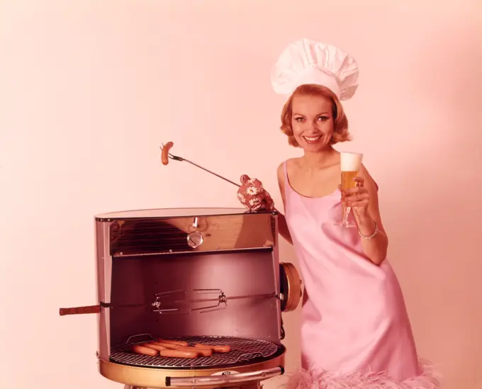 1960S Smiling Blond Woman Wearing Pink Party Dress And Chef Hat Grilling Hot Dogs Drinking Beer