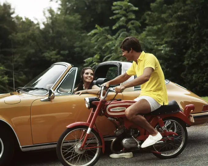 1970S Lifestyle Talking Flirt Couple Man On Small Red Harley Davidson Motorcycle Smiling Woman In Orange Porsche Automobile