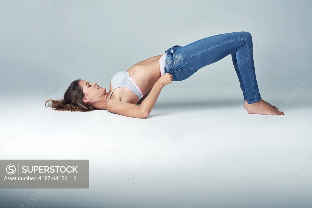 Studio, fashion and woman with jeans for getting ready, dressing and challenge on grey background. Female person, denim and tight pants with struggle for weight gain, diet and stretching on floor