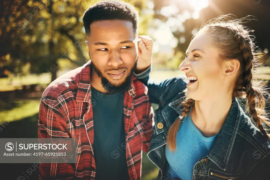 Interracial couple, laughing and smile in nature for playful love, bonding and funny relationship. Happy woman holding man ear with laugh for fun loving care, affection or playing in a park outdoors