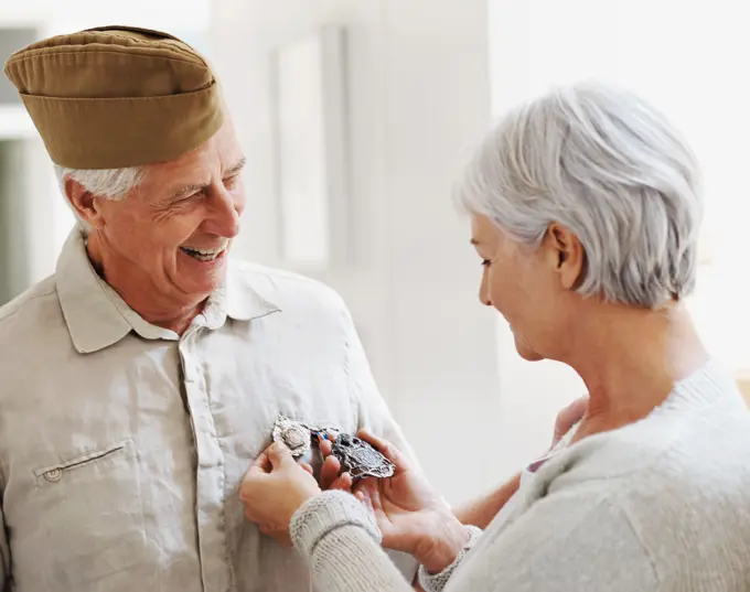 Military veteran, man and woman with medal, uniform and smile together with memory, pride and success. Elderly couple, army badge or regalia with happiness, check and retirement from service in house