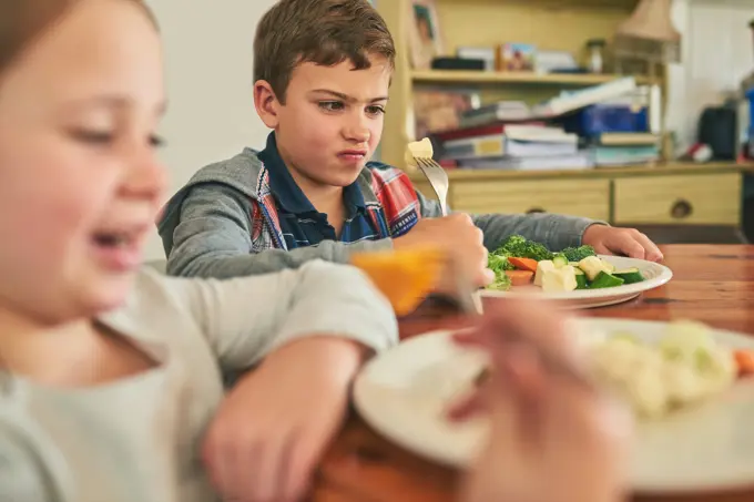 Child, eating and upset with healthy food in home with vegetables, nutrition and angry with diet. Hungry, kid and frustrated with plate of potatoes, salad and dislike for dinner with broccoli