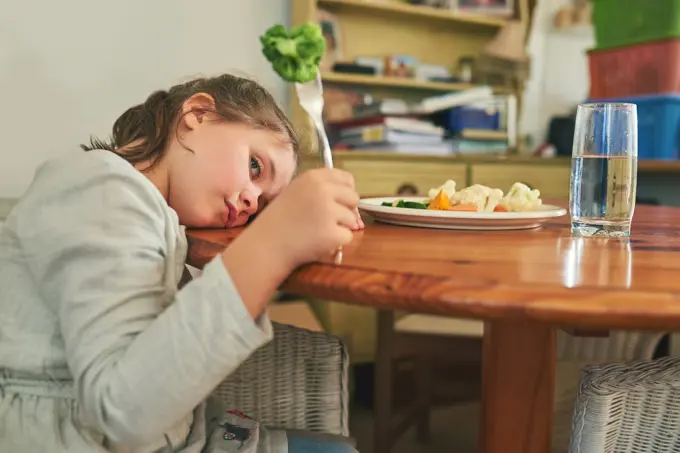 Angry, child and eating healthy food in home with vegetables, nutrition and tired of green diet. Hungry, kid and frustrated with plate of broccoli, salad and dislike for vegan dinner or choice