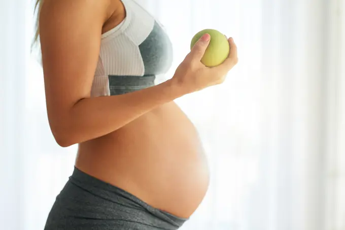 Pregnant, woman and apple for stomach in home, maternity and healthy diet for fetal nutrition. Pregnancy development, prenatal snack and fiber for gestation, obstetrician recommendation for wellness