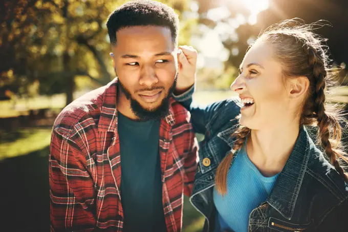 Interracial couple, laughing and smile in nature for playful love, bonding and funny relationship. Happy woman holding man ear with laugh for fun loving care, affection or playing in a park outdoors