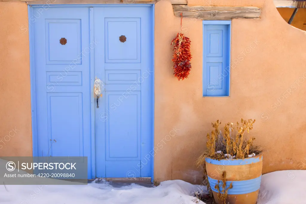 Adobe style house - simple beige coloured house in adobe style with a bright blue coloured door and a string of dried chilly peppers, called ristra, hanging from a beam. In winter - Rancho de Taos, New Mexico, USA .