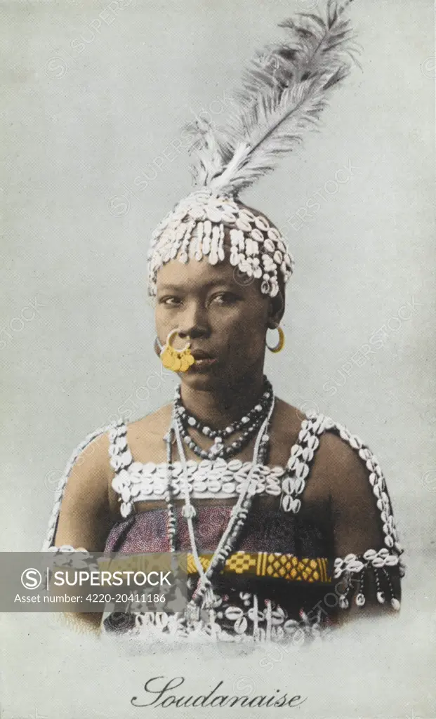 Sudanese Woman, wearing a fantastic decorated nosering, headdress made out of small white cowrie (cowry) shells and topped with large feathers. Her dress is also edged with small shells and beads.     Date: 1910s