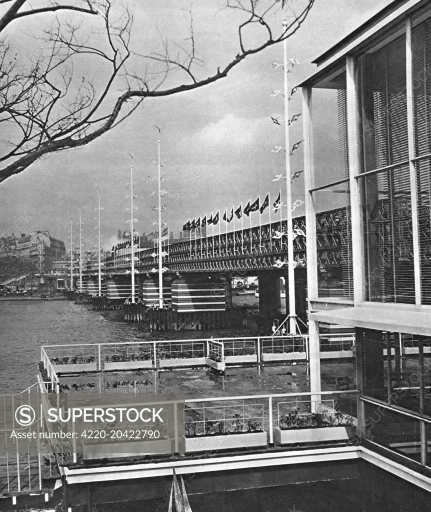 View of the Bailey Bridge across the River Thames, part of the Festival of Britain site, South Bank, London.  The bridge, decorated with flags and wind-driven spinners, links Charing Cross on the north bank with the festival site on the south bank of the river.  The building in the foreground is the two-storey Regatta Restaurant, giving wonderful views of the river.      Date: early 1951