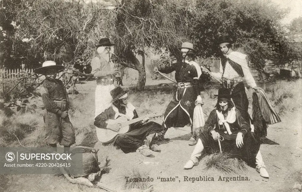 Young Argentinian men in traditional costume drinking Mate, a traditional South American infused drink, prepared from steeping dried leaves of yerba mate (llex paraguariensis, known in Portuguese as erva mate) in hot water. Mate is served with a metal straw from a shared hollow calabash gourd (held in this shot by the gentleman second from left at rear). The other men can be seen holding their bomba/bombilla or drinking straws.     Date: circa 1910s