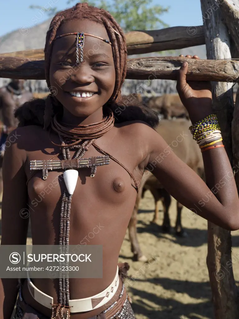 A happy young Himba girl at the entrance to her familys stock pen. Her body gleams from a mixture of red ochre, butterfat and herbs. Her long hair is styled in the traditional Himba way.The Himba are Herero speaking Bantu nomads who live in the harsh, dry but starkly beautiful landscape of remote northwest Namibia.