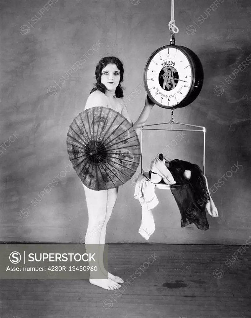 Portrait of a young woman standing in front of a weighing scale and hiding herself with a parasol All persons depicted are not longer living and no es...