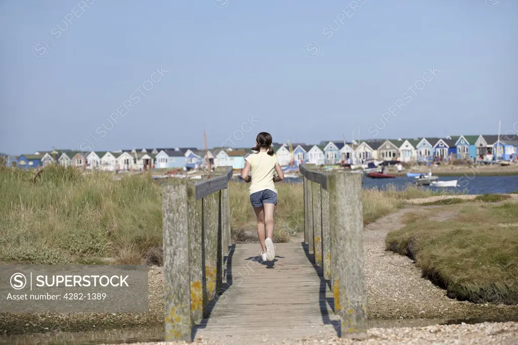 England, Dorset, Christchurch. A young girl running over a foot bridge with beach huts in the background at Christchurch in Dorset.