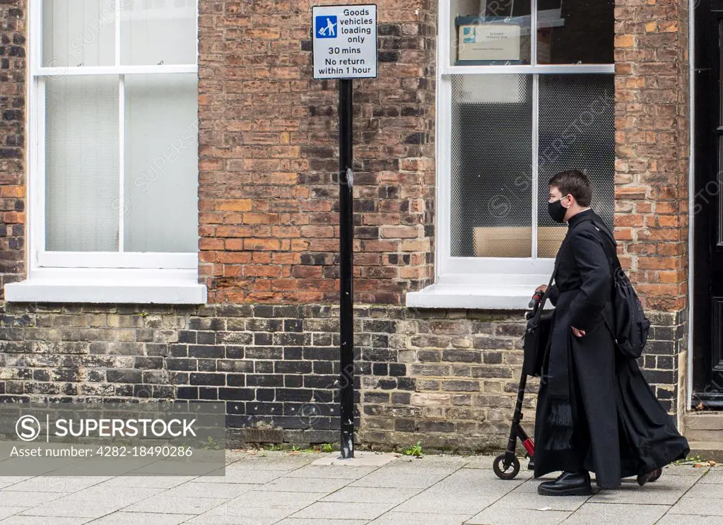 Young priest wearing black robes and face mask and carrying a scooter in Kings Lynn in England.