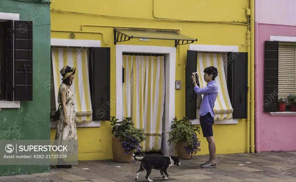 A young tourist has her photo taken by her boyfriend against a colourful background while a dog passes by.