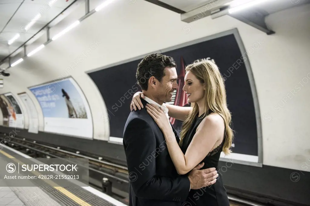 A smartly dressed couple standing cuddling on a platform on the London Underground.