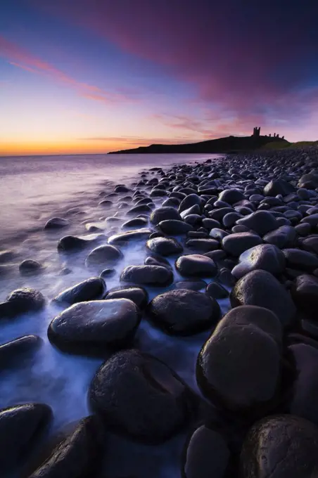 England, Northumberland, Embleton Bay. Waves crash againt the sea sculptured rocks dominating the coastline of Embleton Bay, overlooked by the dramatic ruins of Dunstanburgh Castle.