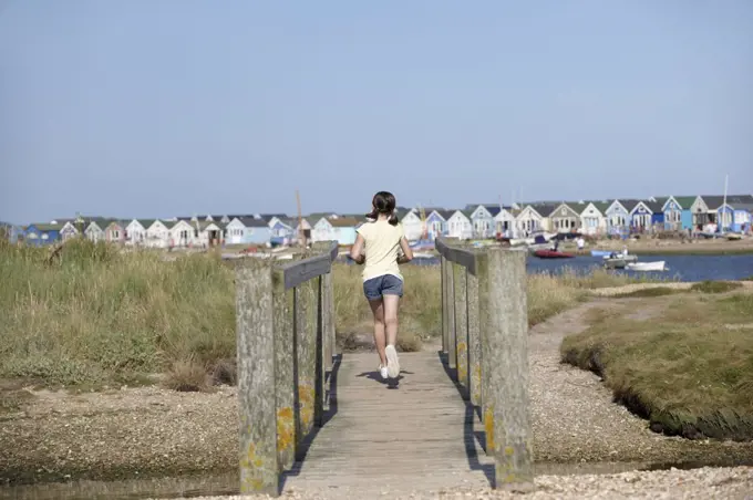 England, Dorset, Christchurch. A young girl running over a foot bridge with beach huts in the background at Christchurch in Dorset.