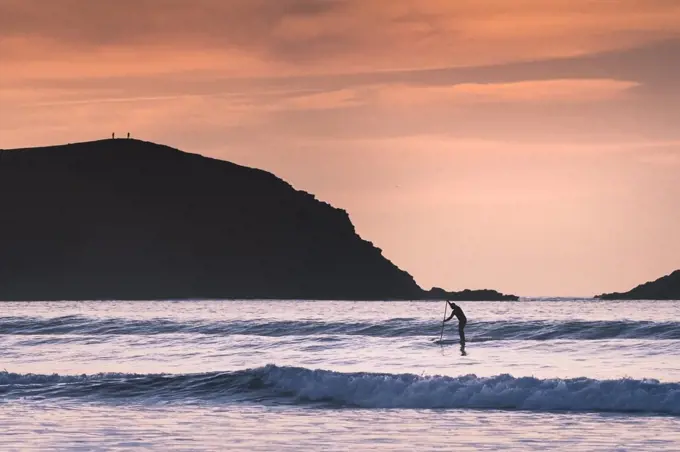 A paddle boarder paddling past Pentire Point East silhouetted by the late evening light at Fistral in Newquay in Cornwall.