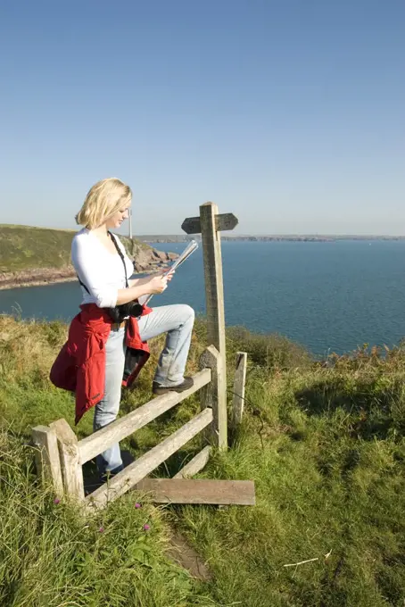 Wales, Pembrokeshire, St. Ann's Head. A female walker stops at a stile to read a map above St. Ann's Head on the Pembrokeshire coastline.