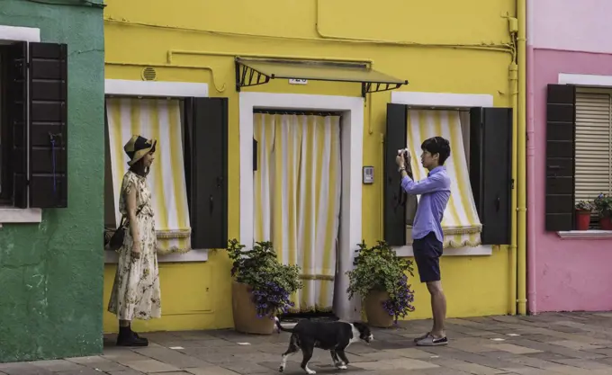 A young tourist has her photo taken by her boyfriend against a colourful background while a dog passes by.