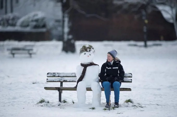 England, London, Wimbledon. Winter day with a woman walker sitting beside a snowman on Wimbledon Common.