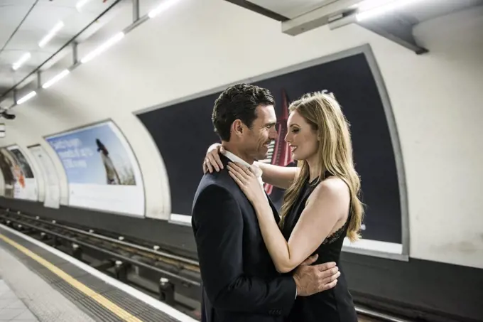 A smartly dressed couple standing cuddling on a platform on the London Underground.