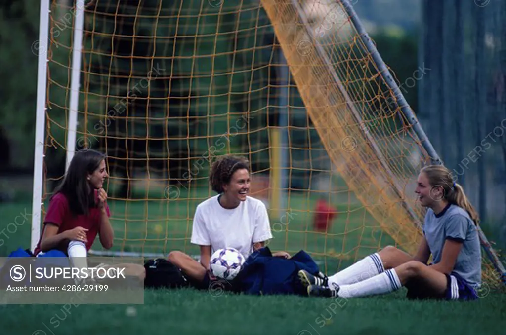 Three teenage soccer girls sitting happily together beside the goalies net.