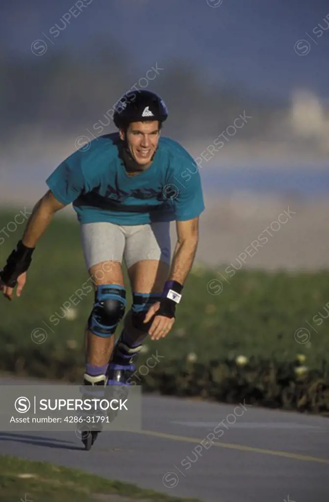 Man wearing a helmet and protective pads rollerblading down a sidewalk near a beach.