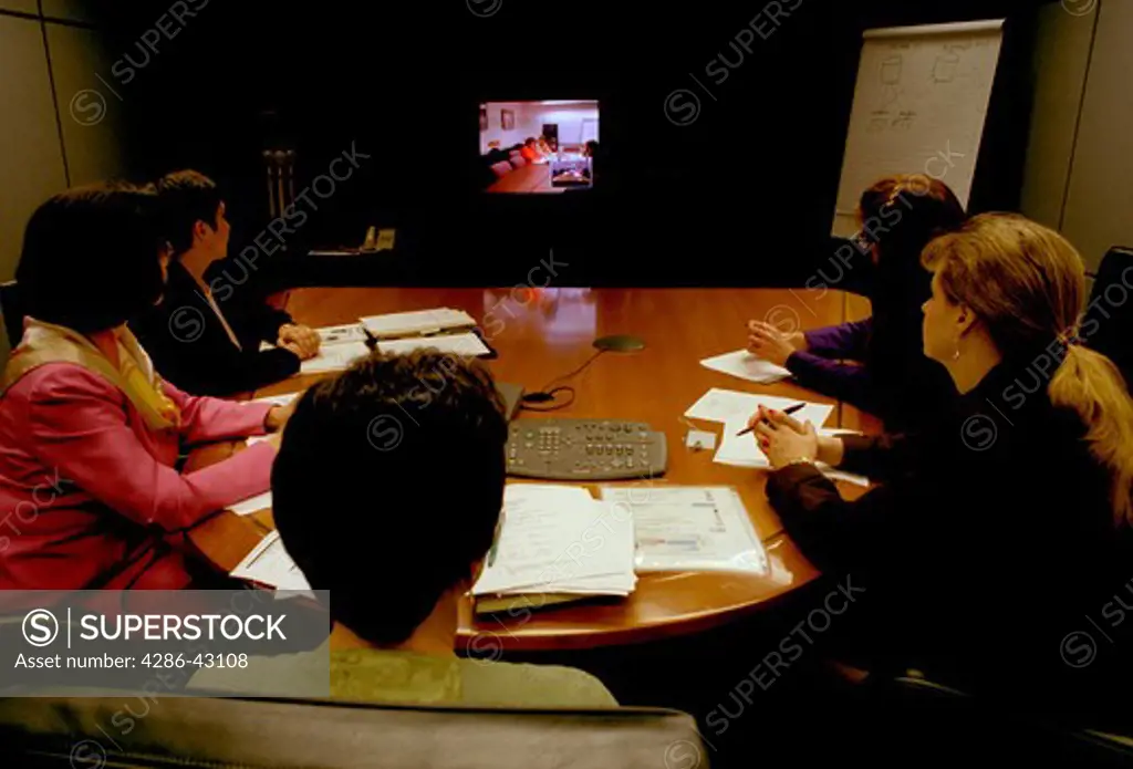 Five businesswomen sitting at a conference table watching a video presentation. 