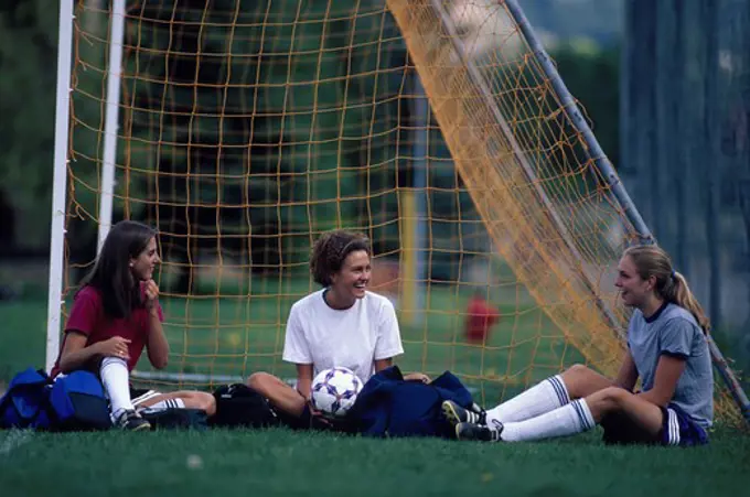 Three teenage soccer girls sitting happily together beside the goalies net.