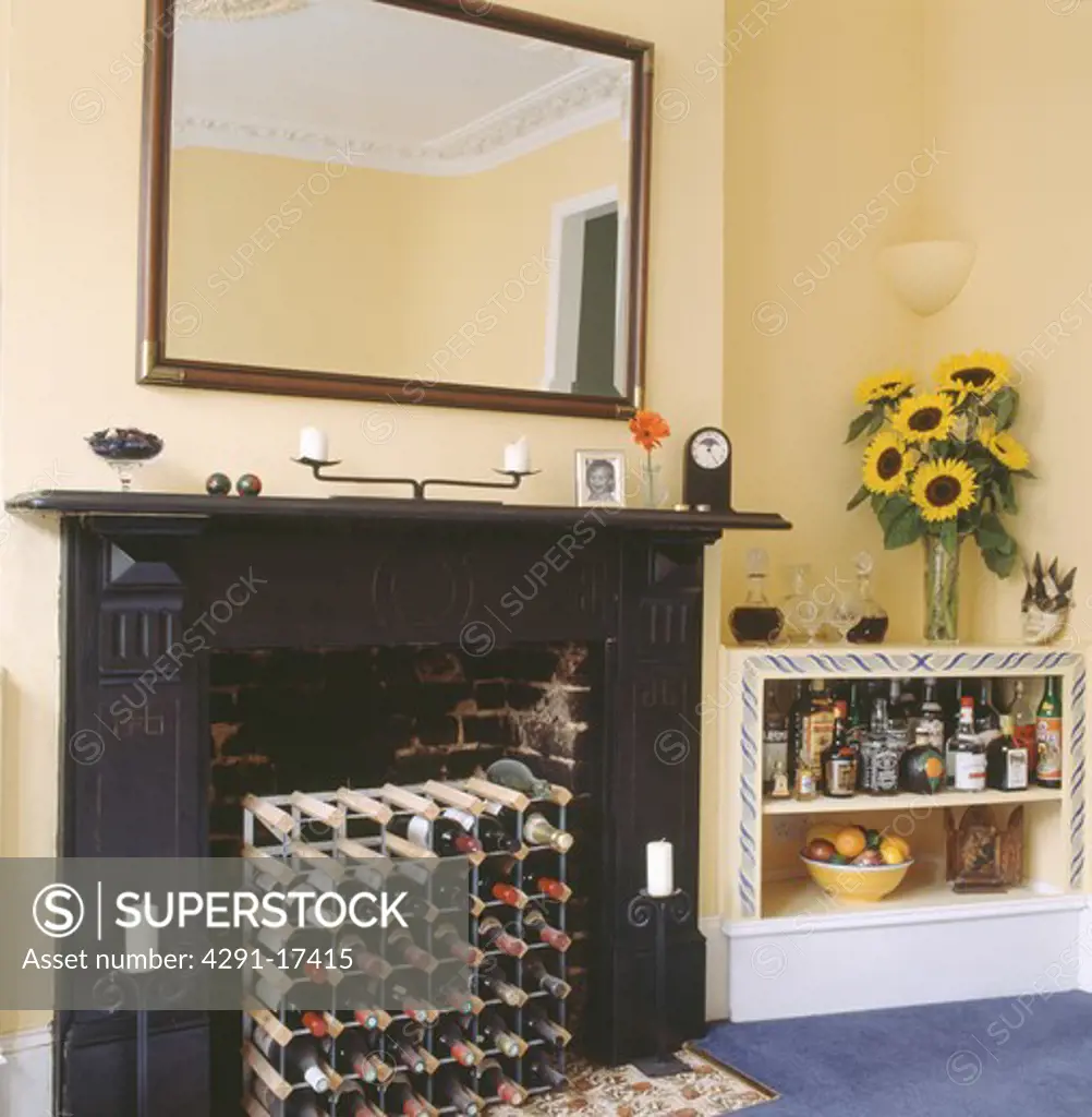 Wine storage in fireplace below large mirror in pastel yellow dining room  with sunflowers and liqueur bottles on shelving - SuperStock